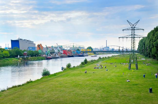 Landschaftsfoto mit Industrieanlage, Fluss und grüner Liegewiese (refer to: For a Systematic Integration of Environment and Health)